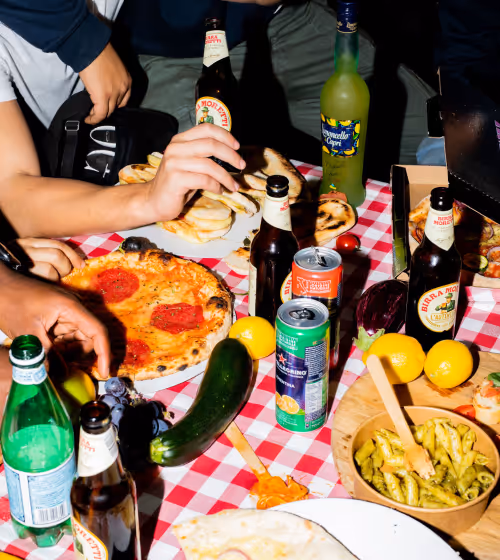 Table with a red and white checkered tablecloth holding pepperoni pizza, bottles of beer, soda cans, lemons, cucumbers, grapes, and bowls of food with people reaching for items.