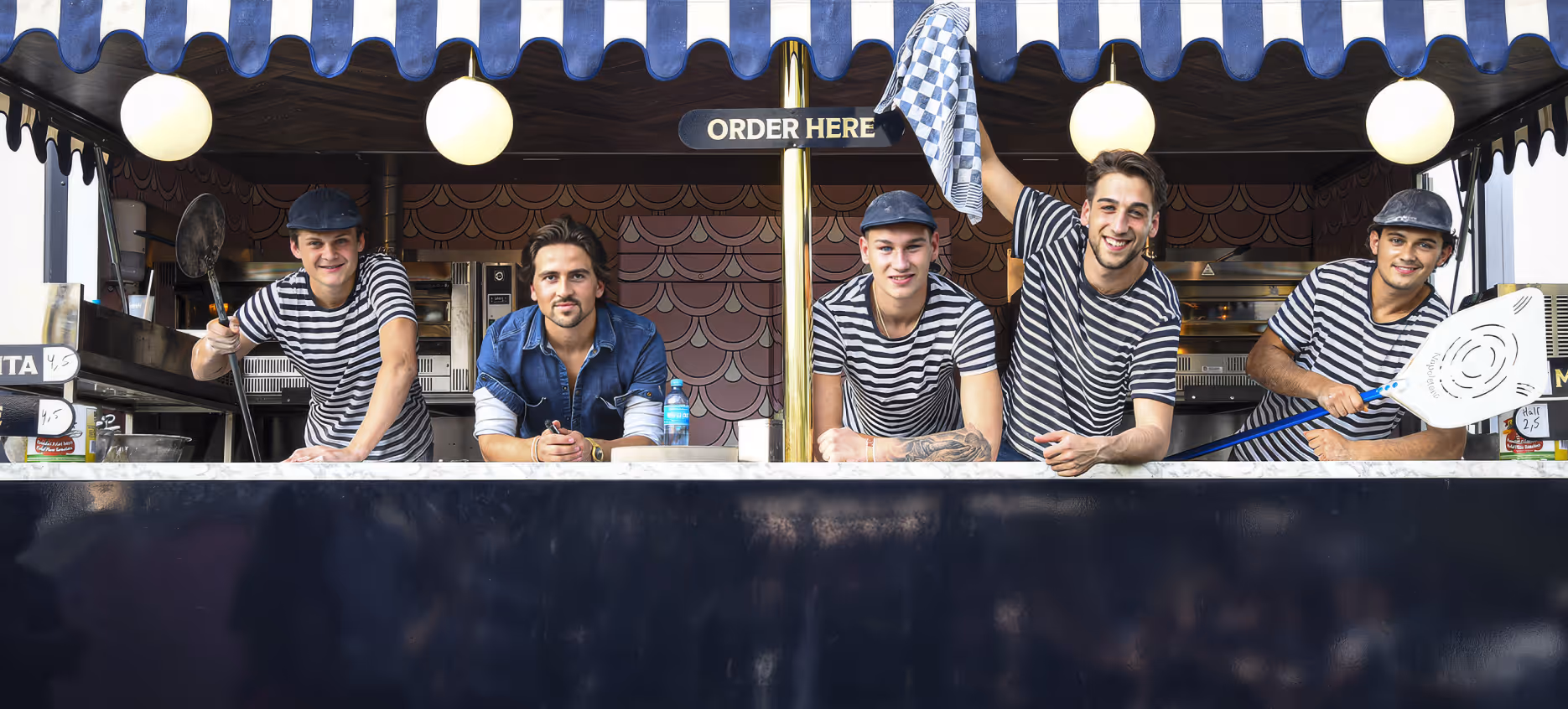 Five smiling men, four wearing striped shirts and two holding pizza peels, standing behind a counter with an 'Order Here' sign above.