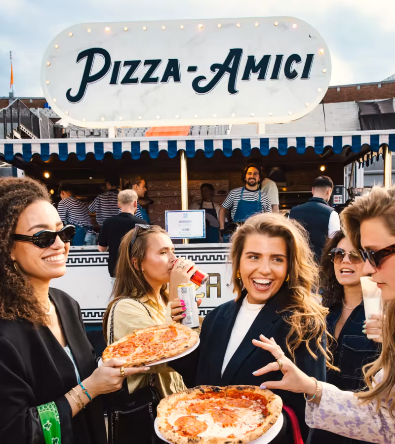 Group of five smiling young people enjoying pizza slices in front of a food stall with a sign reading 'Fornello'.