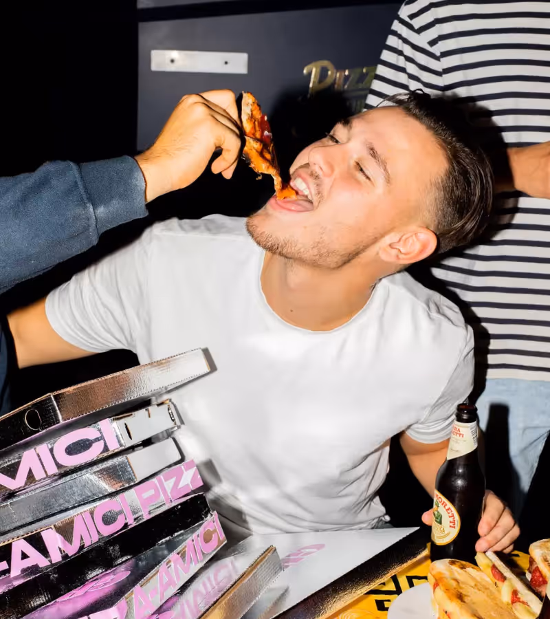 Man in een wit T-shirt die een stuk pizza wordt gevoerd terwijl hij een bierflesje vasthoudt bij een tafel met pizzadozen en broodjes.
