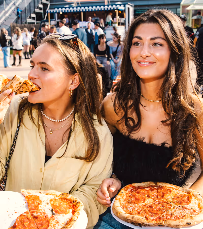 Two young women enjoying pepperoni pizza outdoors at a busy food market.