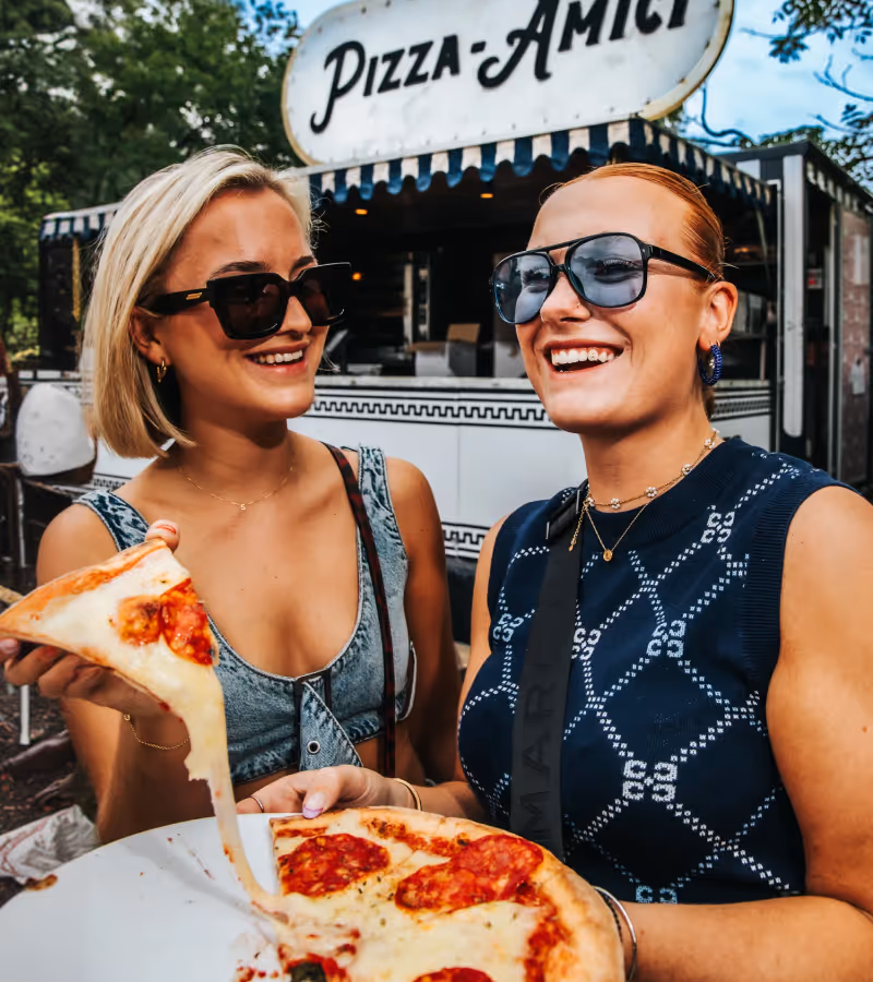 Two smiling women wearing sunglasses holding and sharing a pepperoni pizza slice in front of a food stand with a Fornello sign.