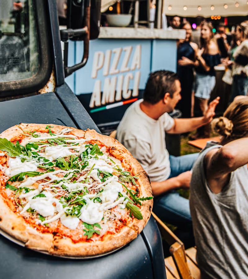 Pizza with arugula and white sauce on a plate resting on a vehicle hood, with people socializing near a food truck labeled Fornello in the background.