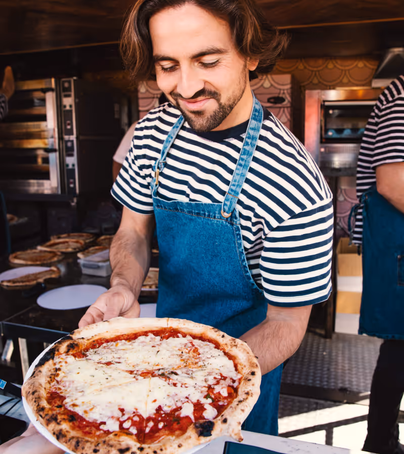 Man with striped shirt and blue apron smiling while handing over a freshly baked cheese pizza.
