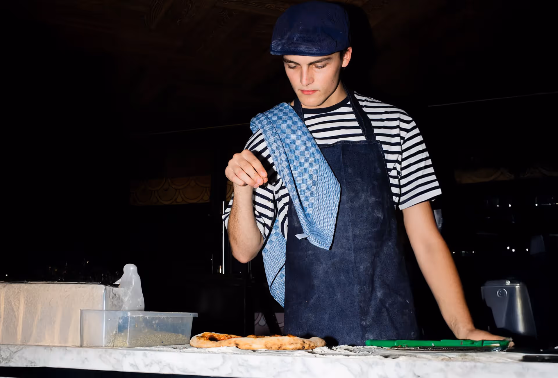 Young male chef wearing a striped shirt and apron with a towel over his shoulder sprinkling seasoning on a pizza base on a marble countertop.