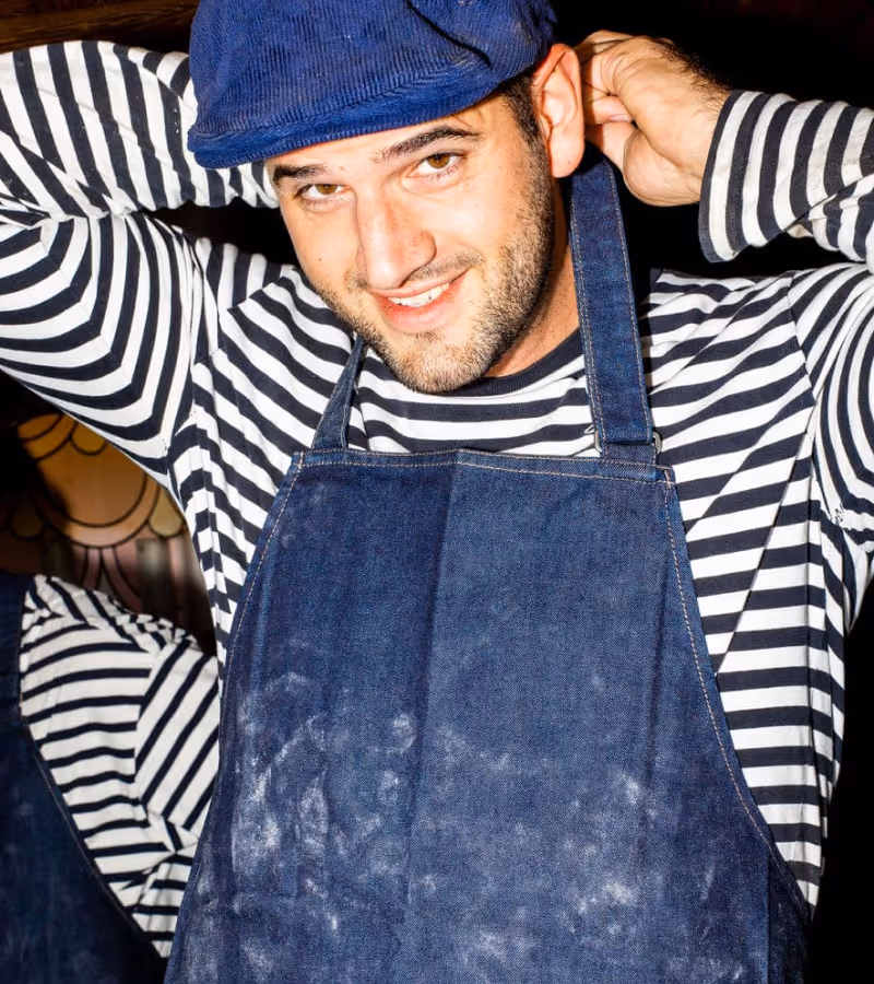 Smiling man wearing a blue cap, striped shirt, and blue apron with flour marks adjusting his cap.