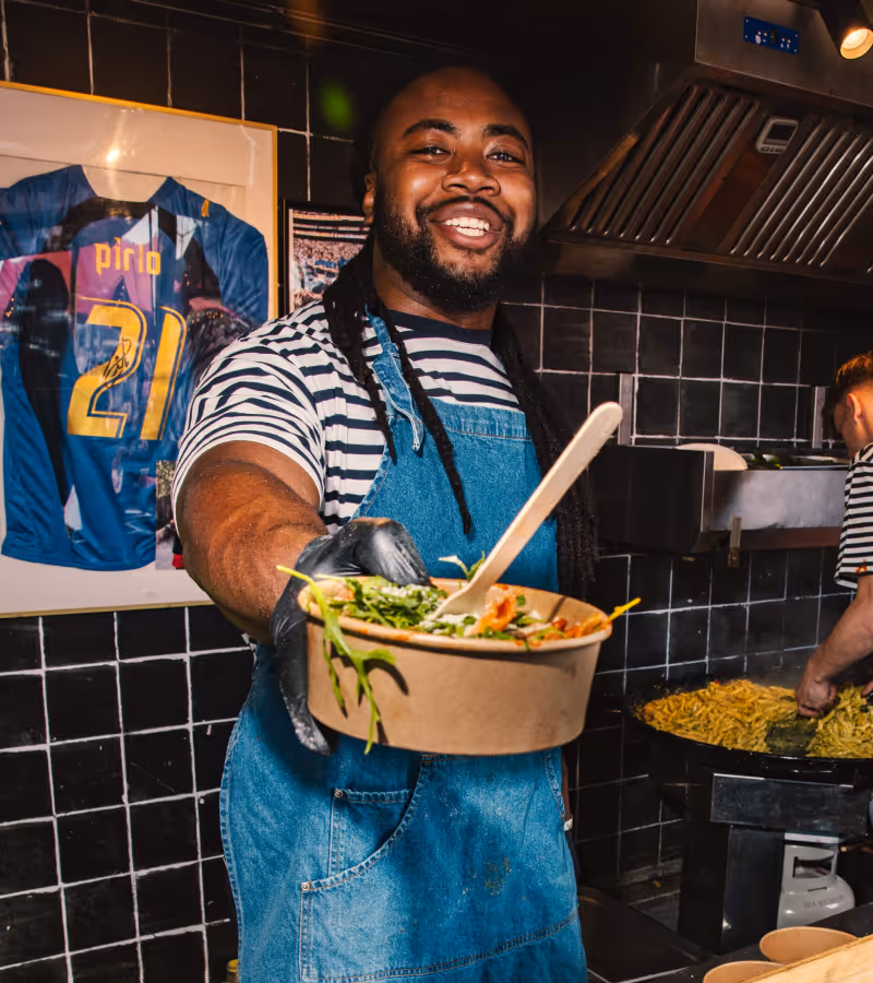 Smiling man in striped shirt and denim apron serving a bowl of fresh salad in a kitchen with black tiled walls.