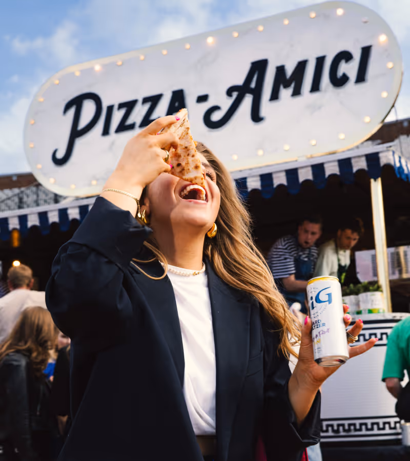 Woman enjoying a slice of pizza and holding a can of hard seltzer in front of a pizza stand named Fornello.