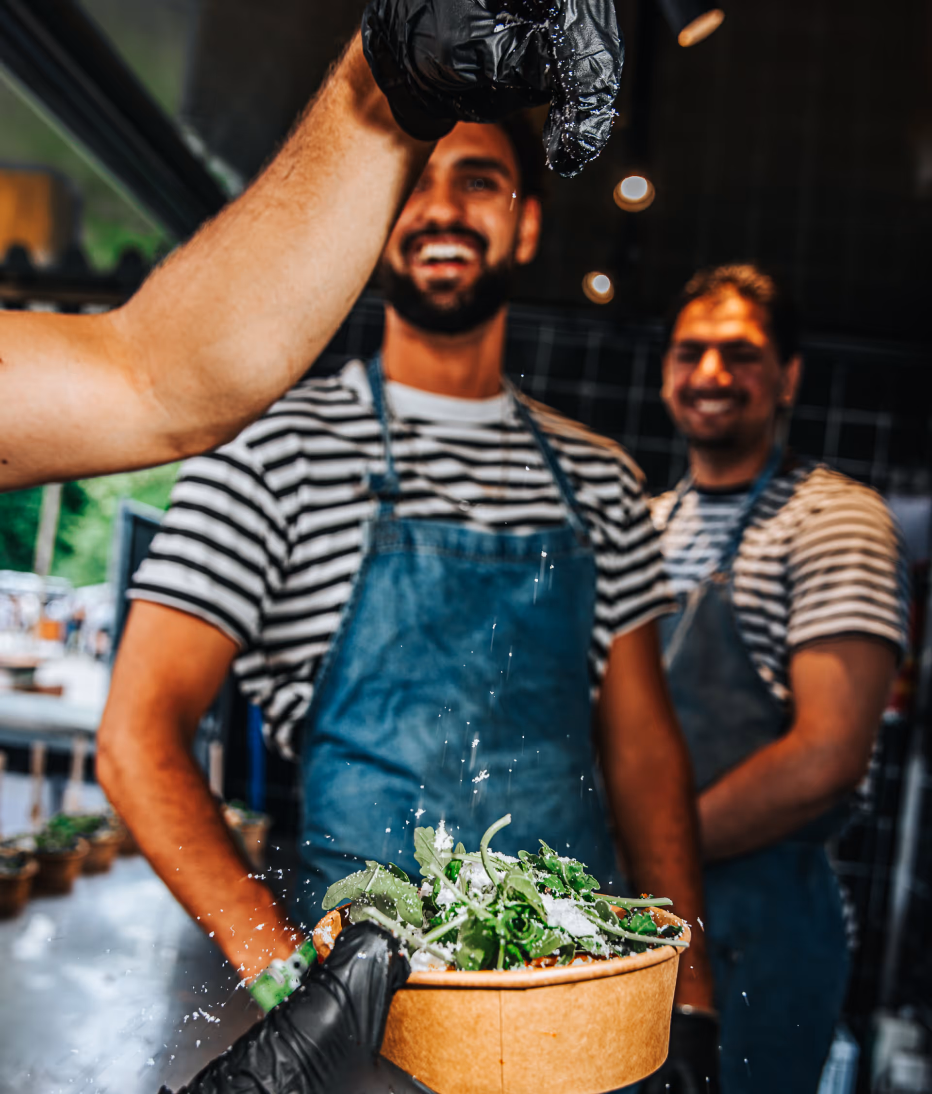 Twee koks in schorten en zwart-witte gestreepte shirts lachen terwijl er een hand met zwarte handschoen ingrediënten over een schaal met sla strooit.