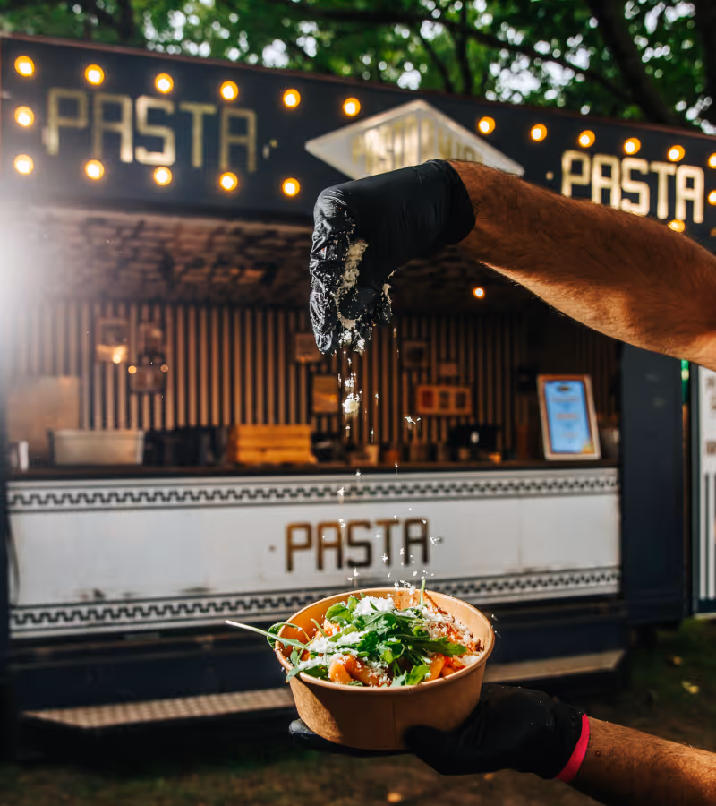 Hand met zwarte handschoen strooit geraspte kaas op een bakje verse pasta met groene kruiden voor een pasta foodtruck.