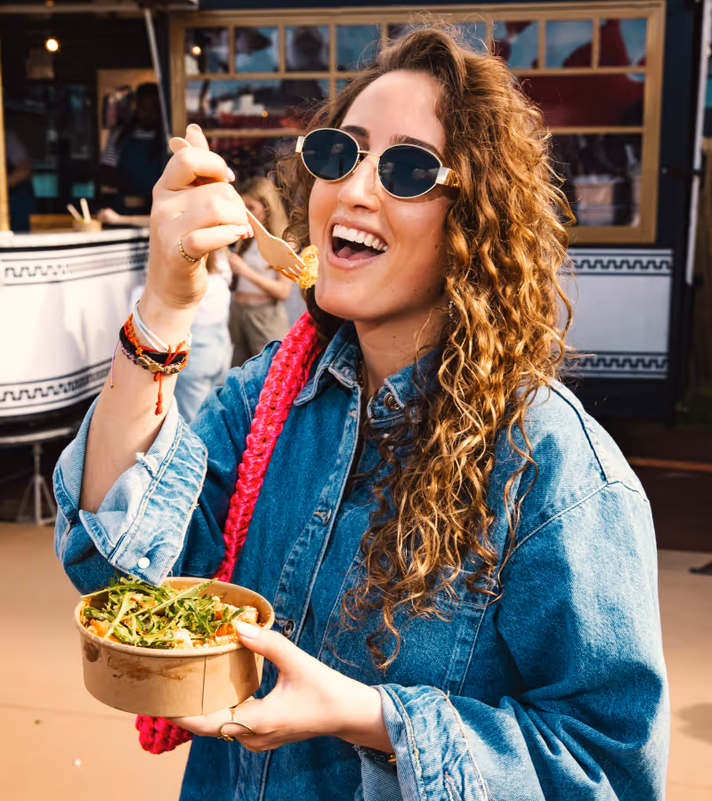 Vrouw met krullend haar en zonnebril die een kom met salade eet, zittend buiten.