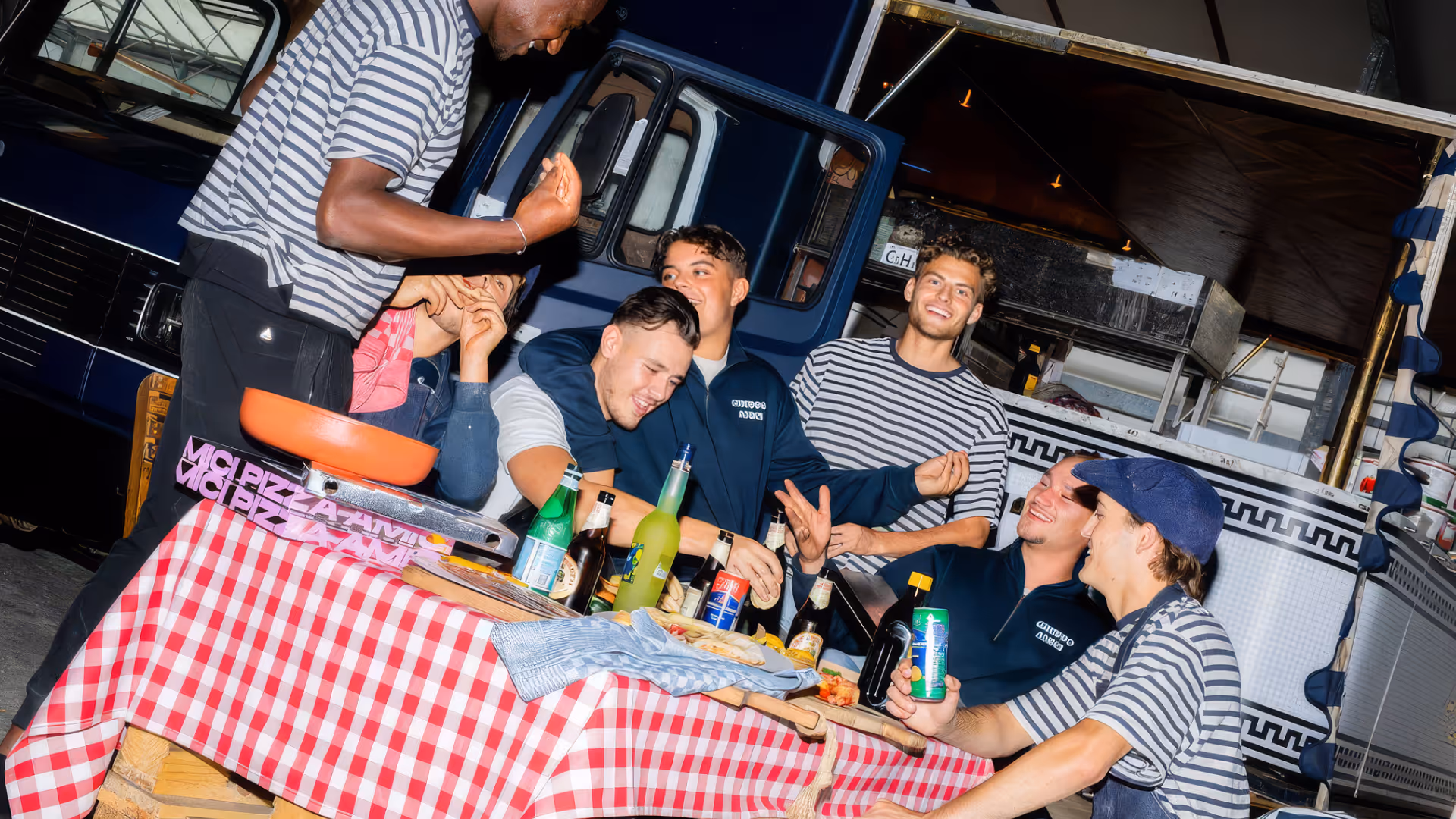 Groep jonge mensen geniet buiten aan tafel met rode geruite tafelkleed, met drankjes en pizza bij een foodtruck in de avond.