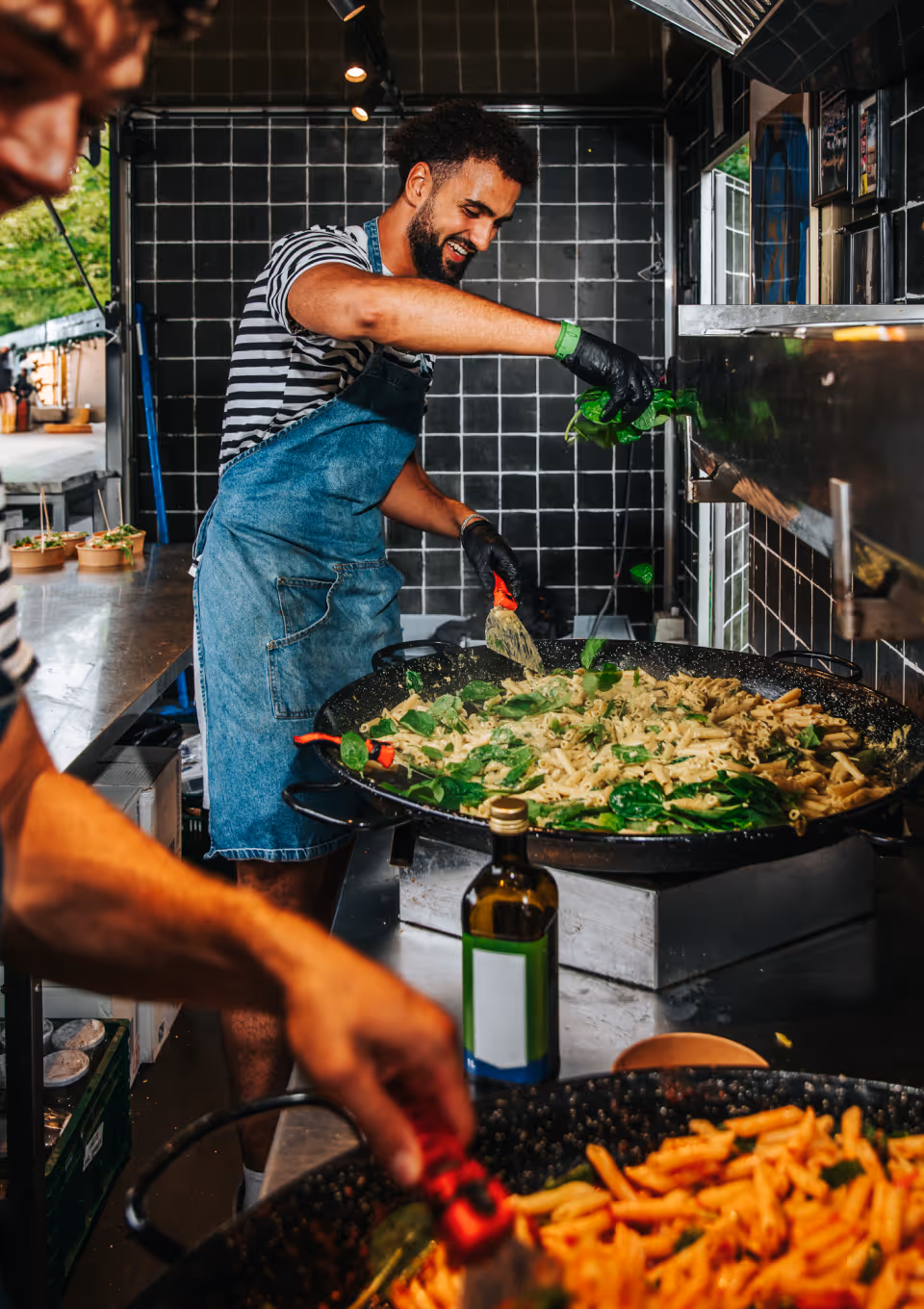 Hand met zwarte handschoen strooit geraspte kaas op een bakje verse pasta met groene kruiden voor een pasta foodtruck.