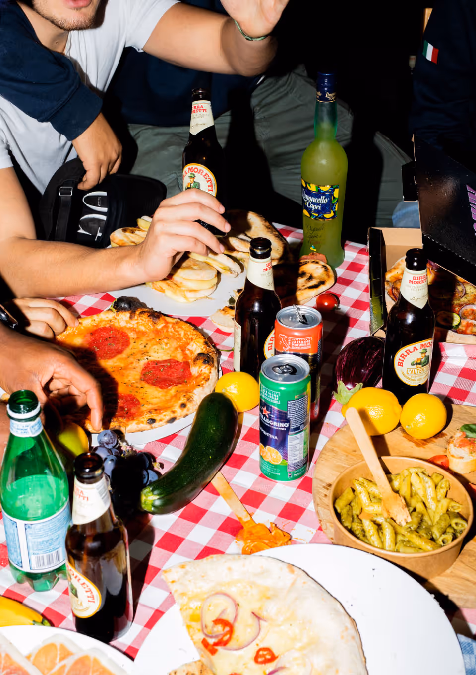 Tafel met diverse Italiaanse gerechten zoals pizza, pasta, brood, citroenen en flessen drank tijdens een samenkomst.