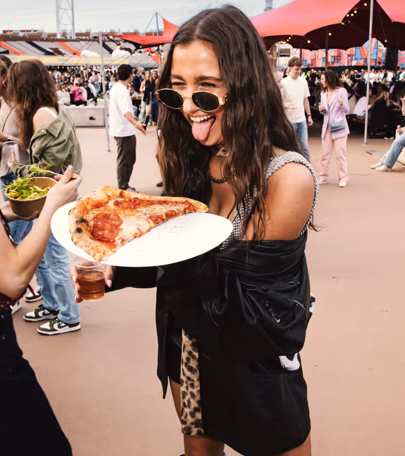 Woman wearing sunglasses playfully sticking out her tongue while holding a plate with a large slice of pepperoni pizza at an outdoor event.