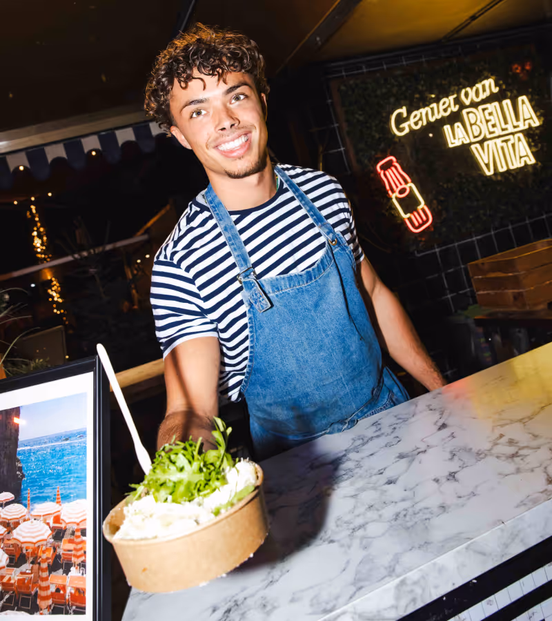 Jonge man met krullend haar en een gestreept shirt draagt een denim schort en biedt een kom salade aan bij een marmeren toonbank in een restaurant.
