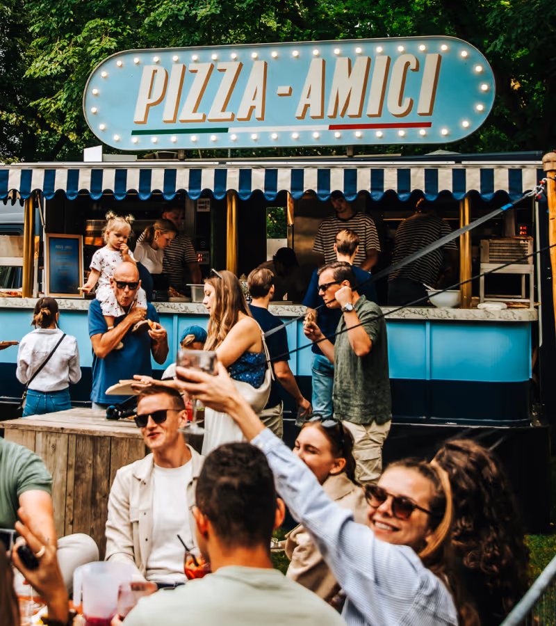 Mensen genieten van een zonnige dag bij een pizza foodtruck met een bord 'PIZZA-AMICI' omlijst met lichtjes.