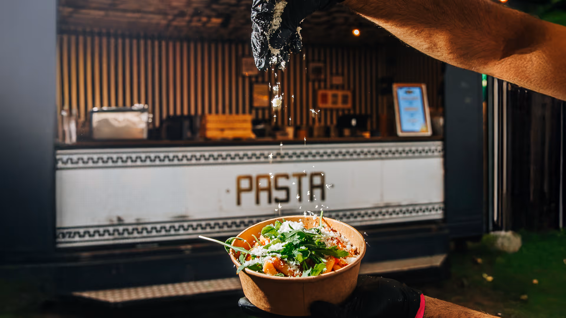 Iemand strooit geraspte kaas over een kom met pasta en verse kruiden in een eetkraam met een bord 'PASTA'.