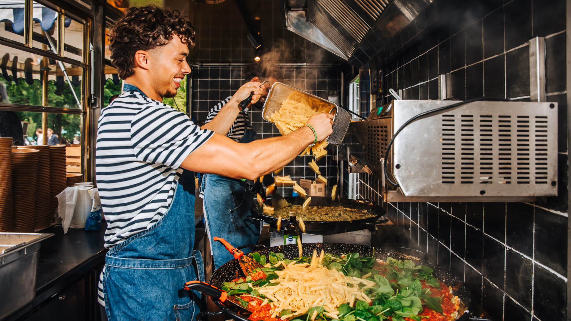 Man met gestreept shirt en spijkerbroek gooit pasta in een grote pan met groenten in een professionele keuken.